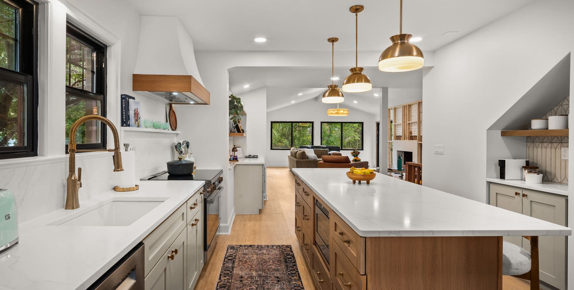 Modern kitchen with white quartz countertops, wood cabinetry, brass pendant lights, and open floor plan connecting to living room with green views through windows