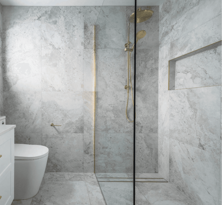 A gray stone bathroom with a walk-in shower with glass partition wall and gold-colored fixtures.