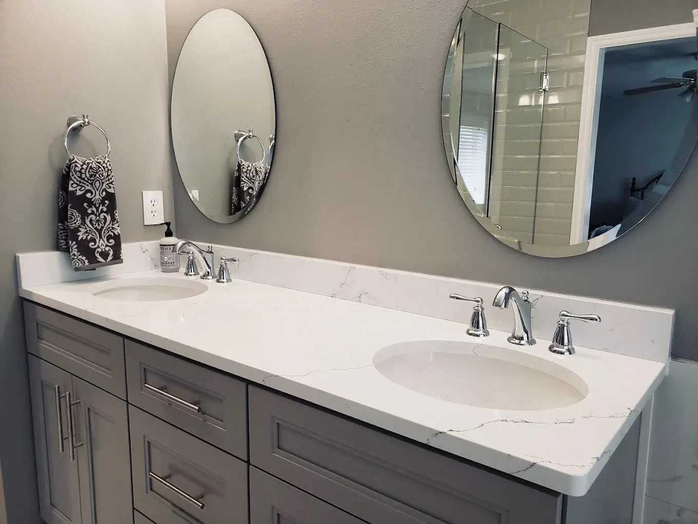 A gray bathroom with double vanity sinks, gray cabinets, white marbled countertop, and two oval-shaped mirrors.