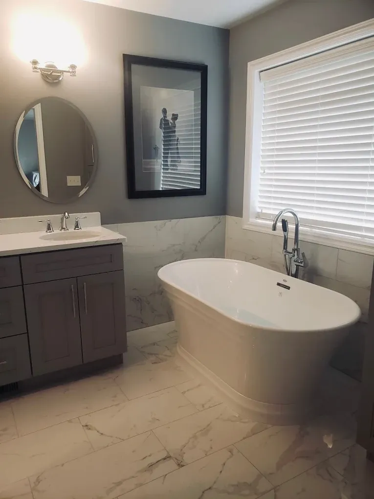 A large soaking bathtub sits in front of a window in a bathroom with white marbled floors.