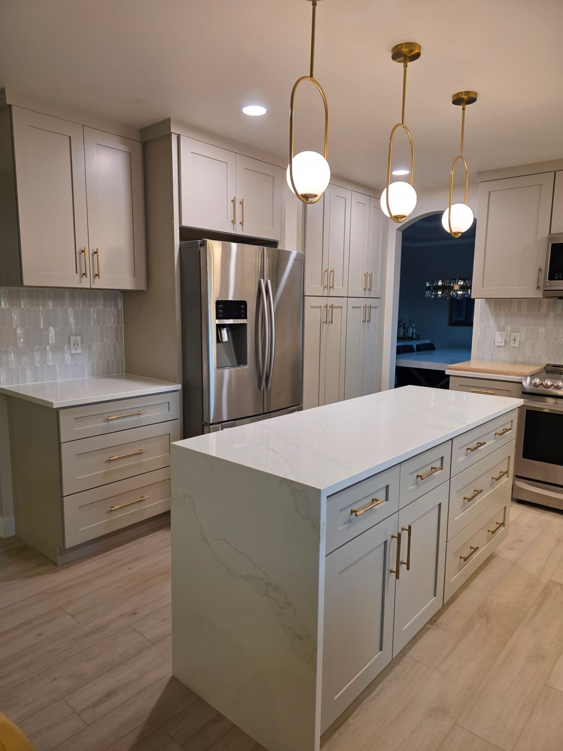 A newly-remodeled kitchen with light gray cabinets, brass-colored fixtures, central island, and new white marbled countertops.