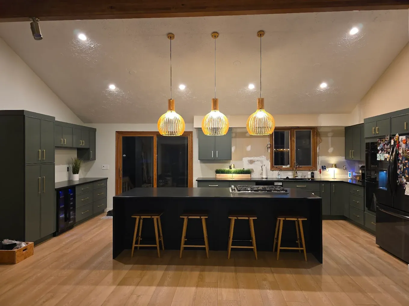 A kitchen with dark gray-green cabinets and a large central island with black stone waterfall countertop and bar stool seating.